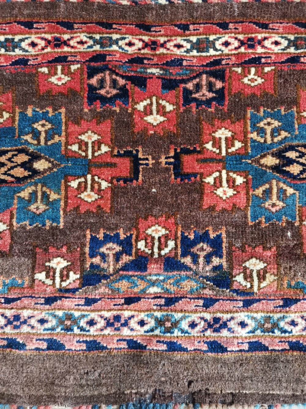 A rectangular hand-knotted wool Yomut torba featuring a dark brown background, geometric red and blue guls, a patterned border, and loose wool fringe along the bottom edge. View II