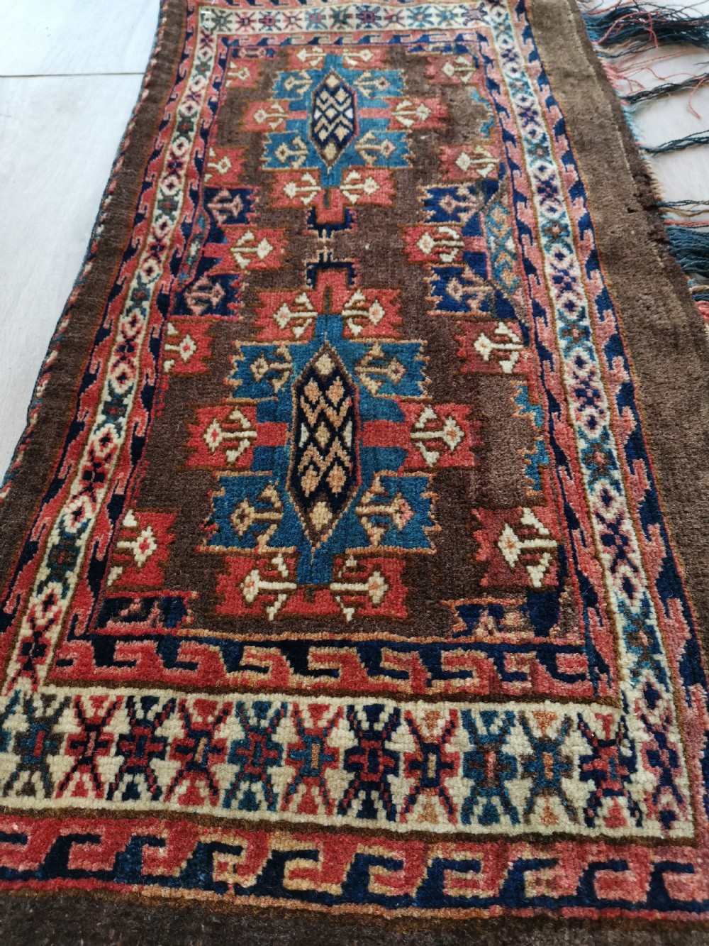 A rectangular hand-knotted wool Yomut torba featuring a dark brown background, geometric red and blue guls, a patterned border, and loose wool fringe along the bottom edge. View III