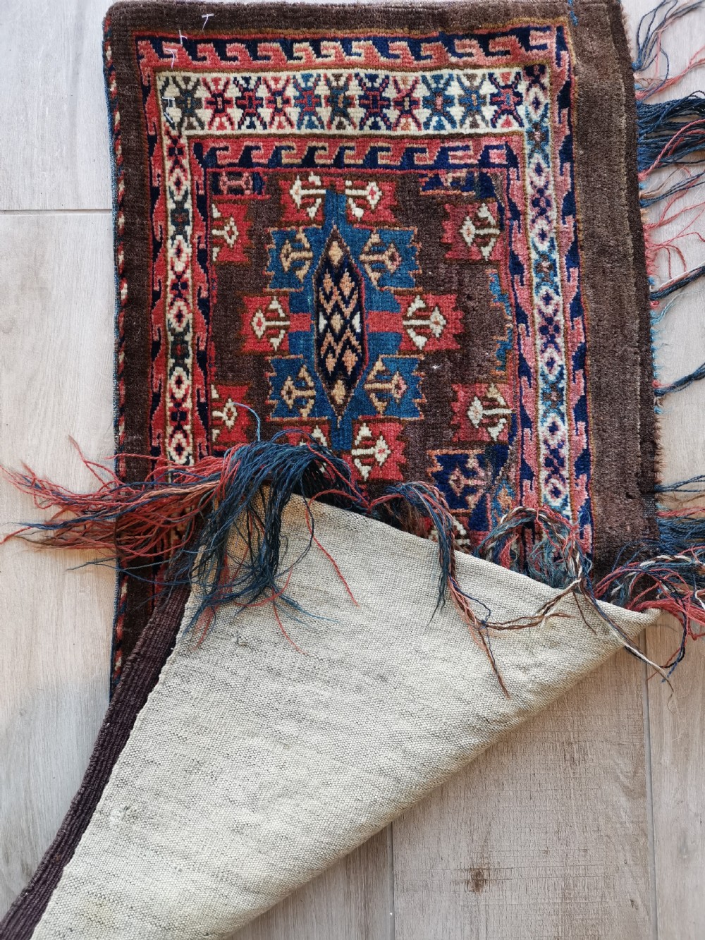 A rectangular hand-knotted wool Yomut torba featuring a dark brown background, geometric red and blue guls, a patterned border, and loose wool fringe along the bottom edge. View IV