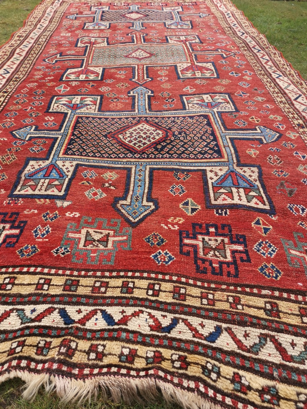 A hand-knotted tribal wool area rug featuring three central geometric stepped medallions in dark blue and ivory on a bold red field surrounded by patterned borders. View II