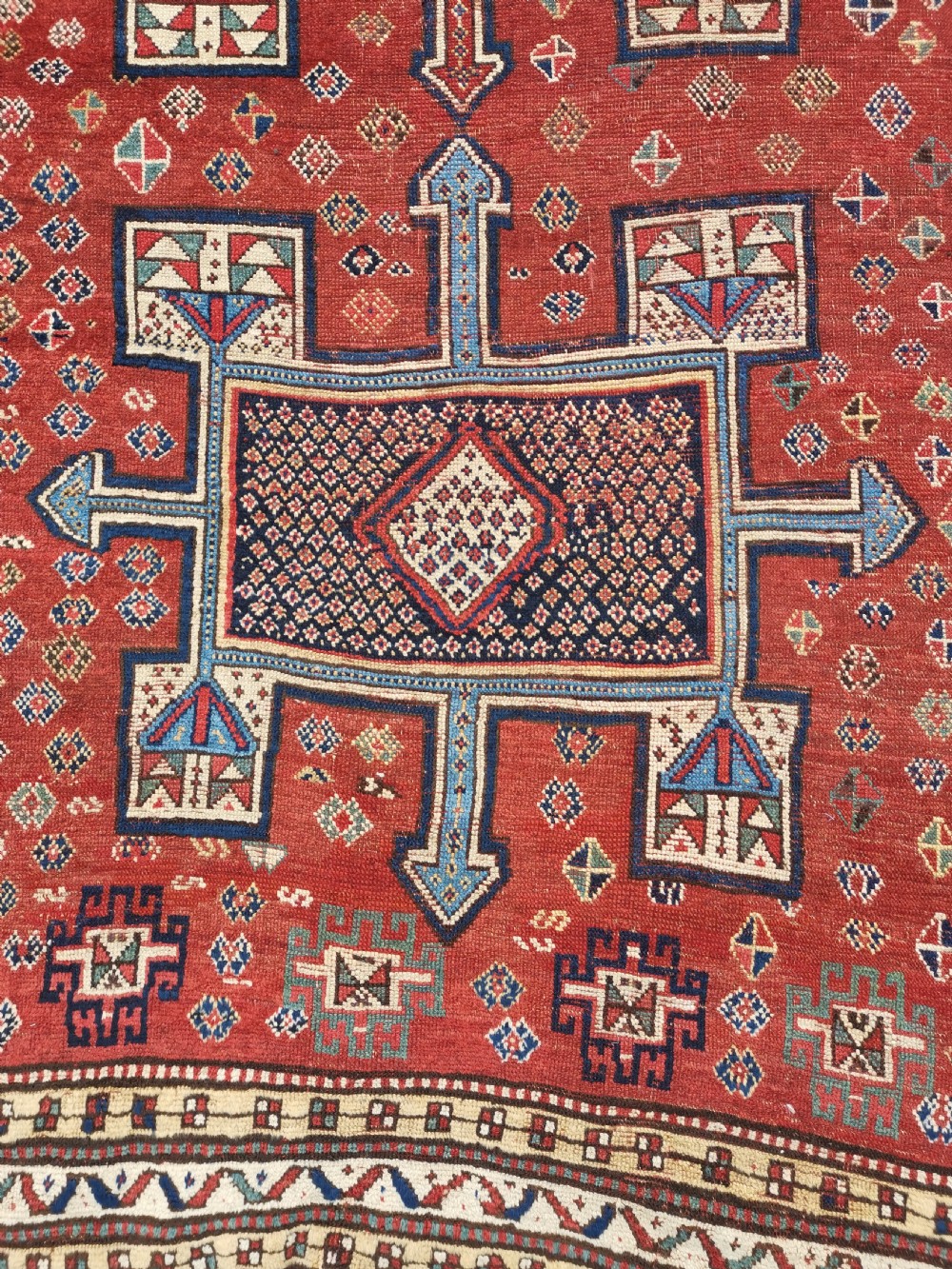 A hand-knotted tribal wool area rug featuring three central geometric stepped medallions in dark blue and ivory on a bold red field surrounded by patterned borders. View III
