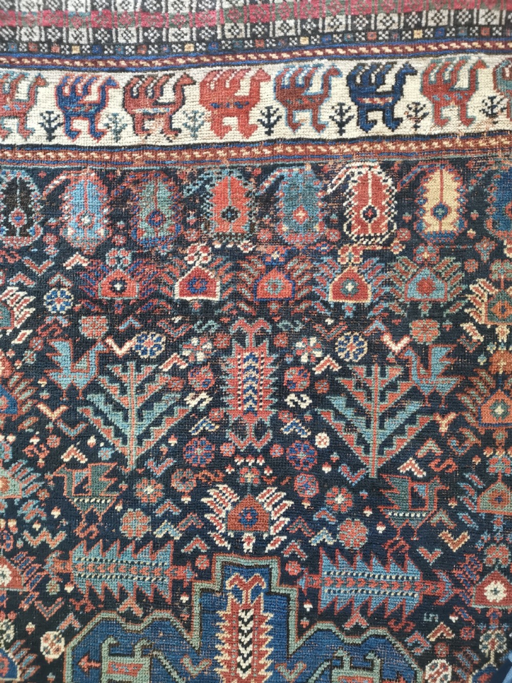 A rectangular hand-knotted wool rug featuring a dark navy central field with a large blue geometric medallion, surrounded by stylised bird motifs and a cream pattern border. View V