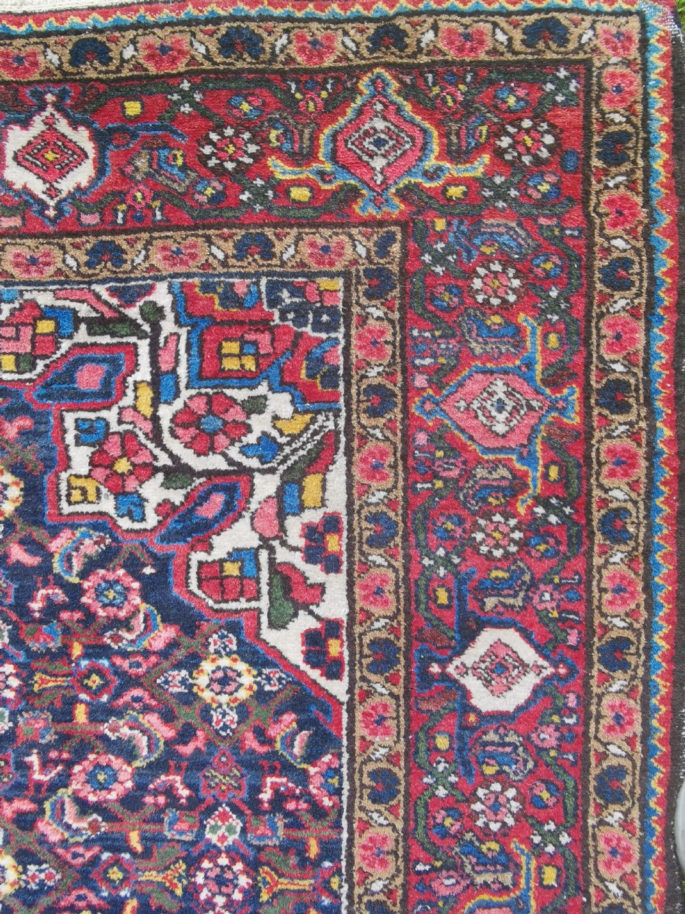 A hand-knotted Persian Hamadan wool rug featuring a central red and blue diamond medallion set on an indigo floral field with a red geometric border. View IV