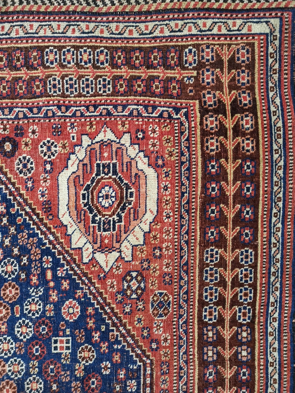 A hand-knotted antique Qashqai wool rug featuring a central indigo diamond medallion surrounded by a terracotta field and geometric borders. View IV