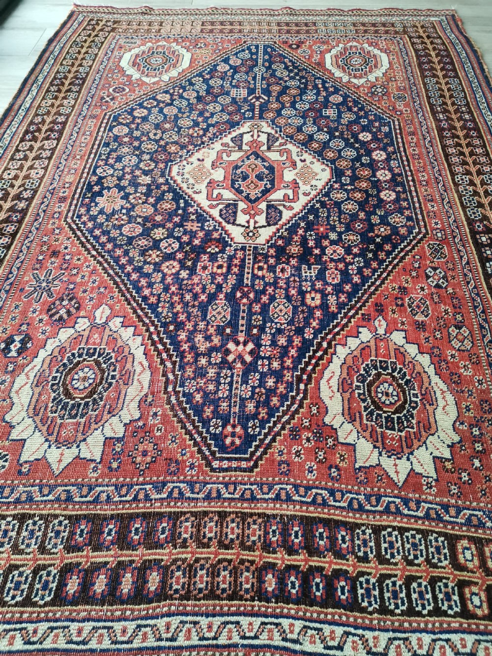 A hand-knotted antique Qashqai wool rug featuring a central indigo diamond medallion surrounded by a terracotta field and geometric borders. View VI
