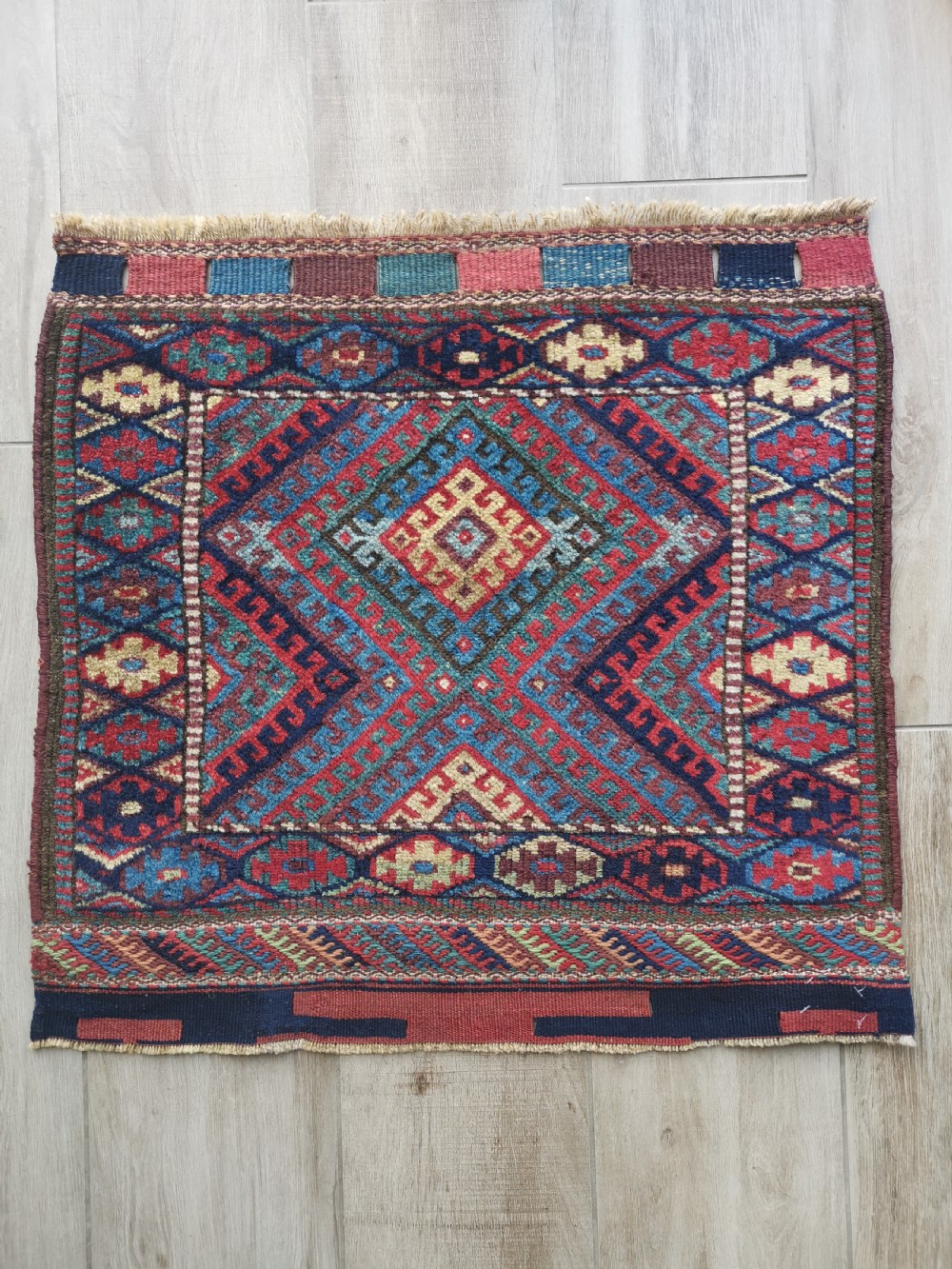 A square handwoven antique Kurdish bagface featuring a central diamond motif surrounded by geometric patterns in red, blue, green, and cream wool.