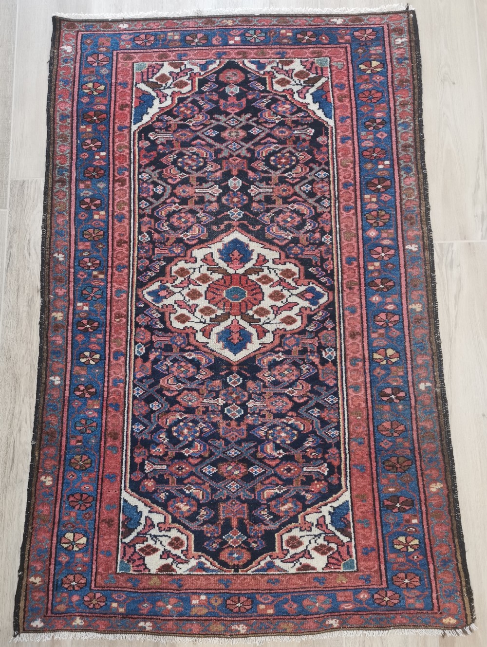 A rectangular hand-knotted wool rug featuring a central cream diamond medallion surrounded by a dark navy field with intricate geometric floral motifs and reddish-pink and blue borders.