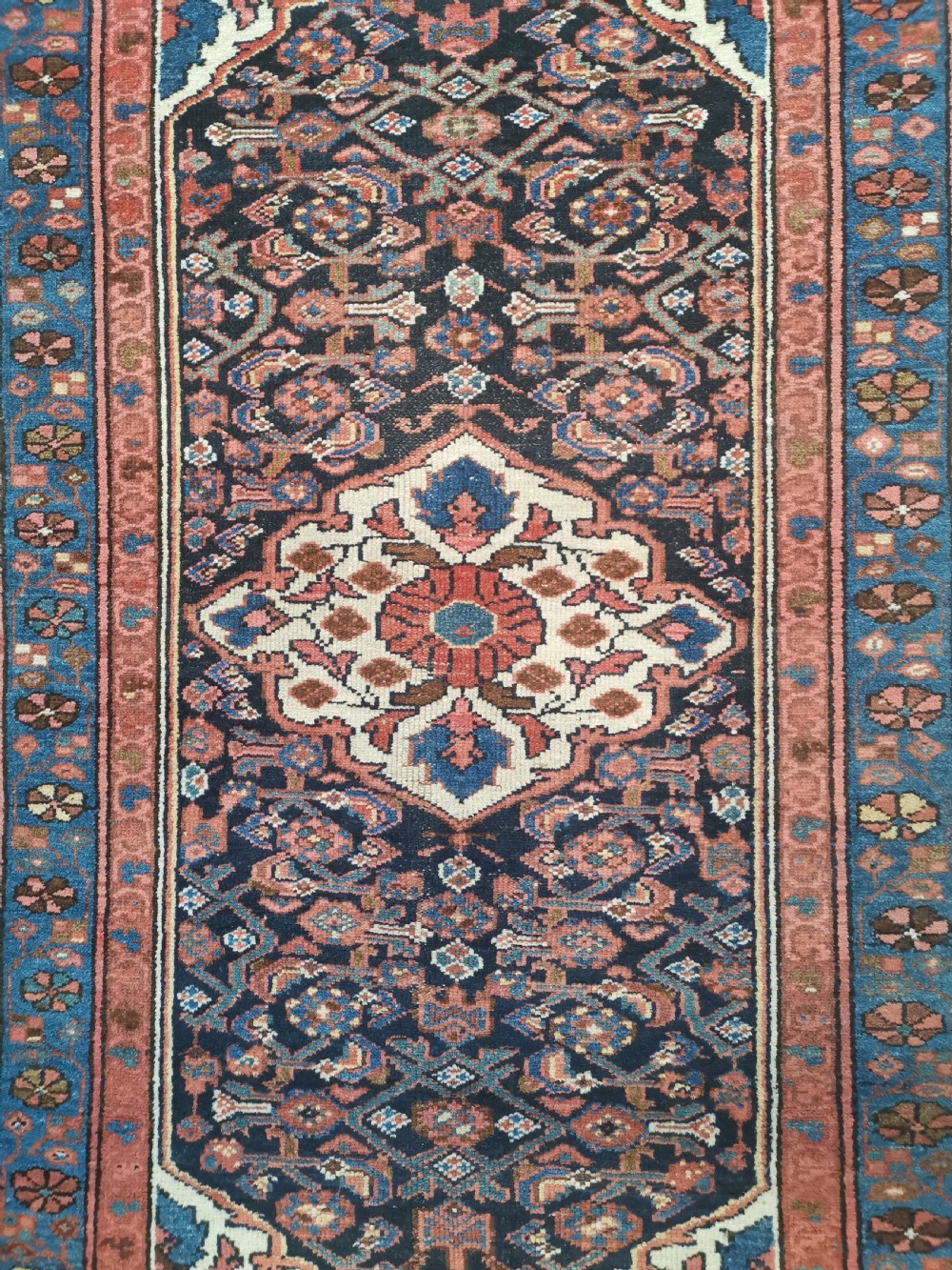 A rectangular hand-knotted wool rug featuring a central cream diamond medallion surrounded by a dark navy field with intricate geometric floral motifs and reddish-pink and blue borders. View III