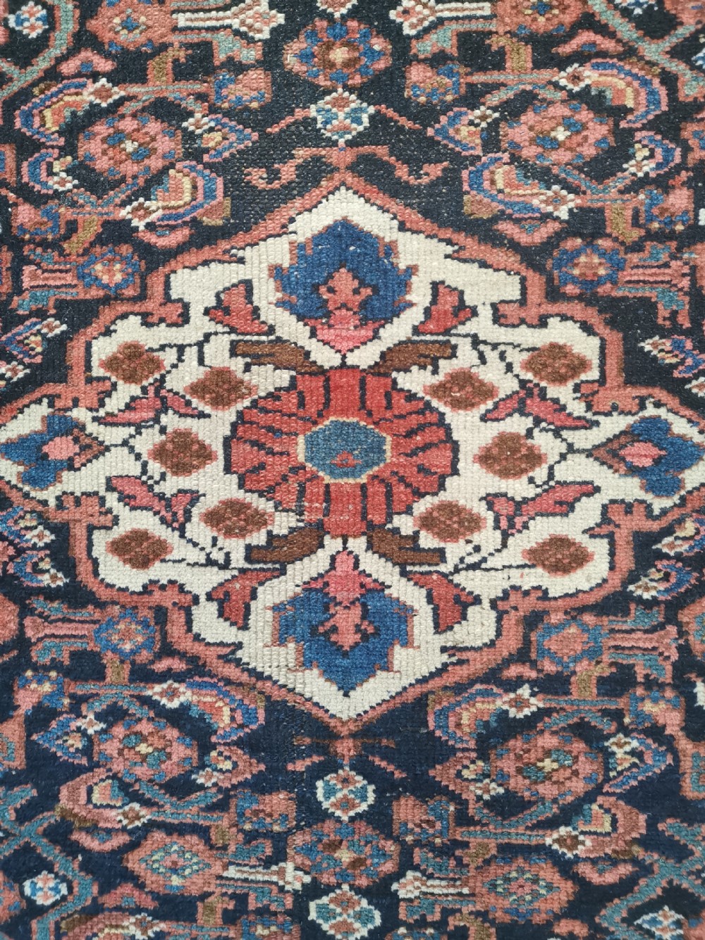 A rectangular hand-knotted wool rug featuring a central cream diamond medallion surrounded by a dark navy field with intricate geometric floral motifs and reddish-pink and blue borders. View IV