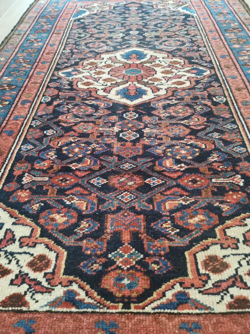 A rectangular hand-knotted wool rug featuring a central cream diamond medallion surrounded by a dark navy field with intricate geometric floral motifs and reddish-pink and blue borders. View V