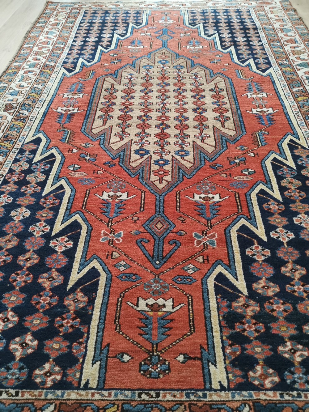 A hand-knotted Mazlaghan wool rug featuring a terracotta stepped central medallion surrounded by a dark blue geometric field and intricate multi-layered borders. View VI