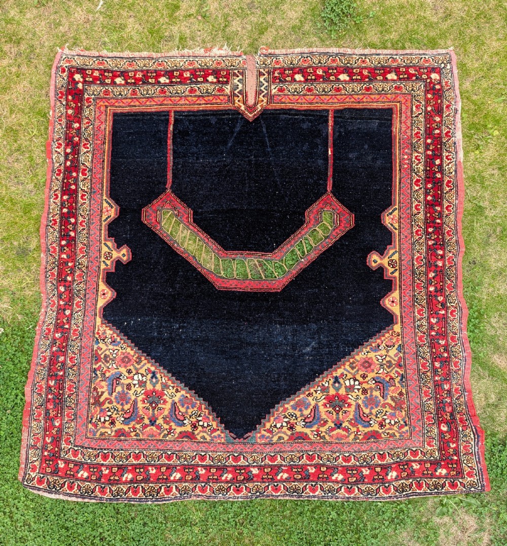 An antique hand-knotted Persian Senneh saddle cover featuring a dark navy field with a central saddle notch, floral spandrels, and multi-banded red patterned borders.