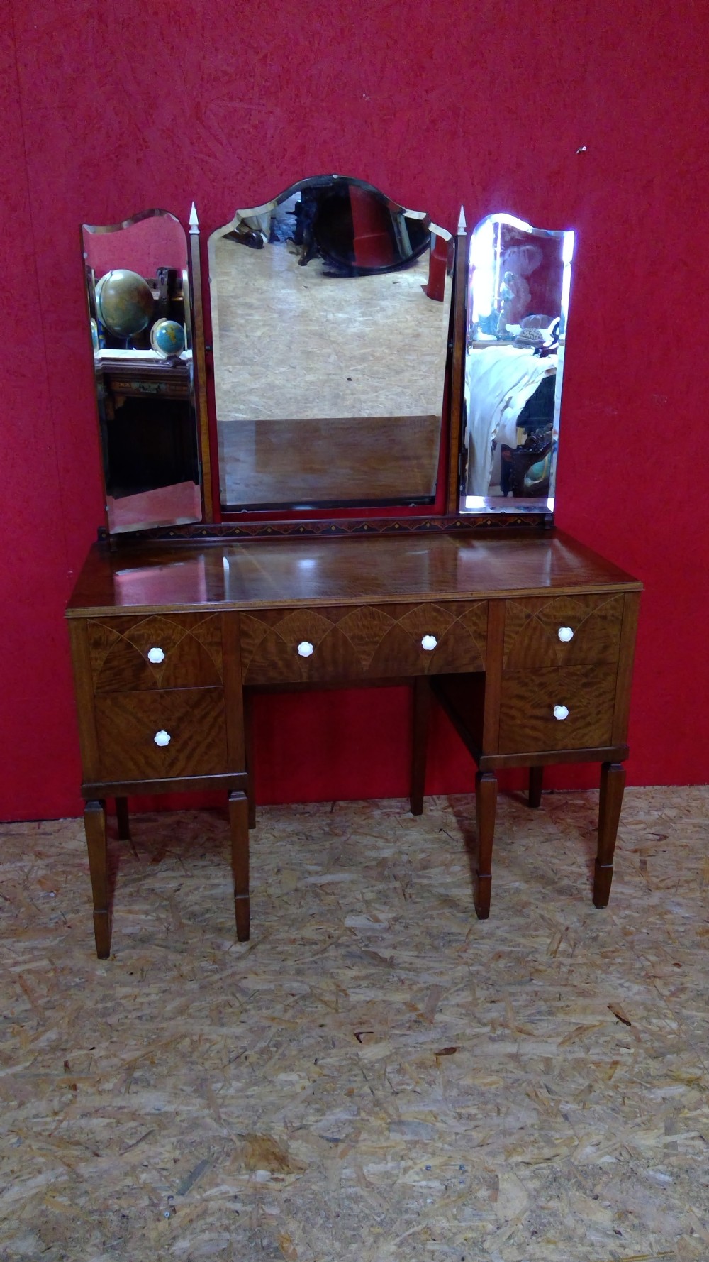An Art Deco mahogany dressing table featuring a central mirror flanked by two adjustable wing mirrors, geometric wood inlays, and small white drawer knobs.