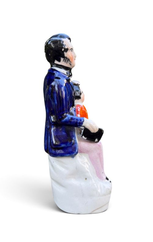 fig 1. An antique Victorian Staffordshire earthenware figure depicting Lord Shaftesbury in a cobalt blue coat seated with a young child in an orange jacket on a white base.