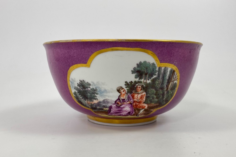 A deep porcelain bowl with a vibrant puce exterior featuring a shaped gilded frame around a hand-painted scene of two figures outdoors.