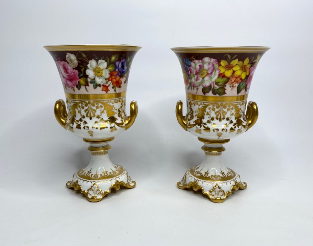 A pair of Royal Crown Derby porcelain urns featuring elaborate gold scrollwork and detailed hand-painted floral bands.