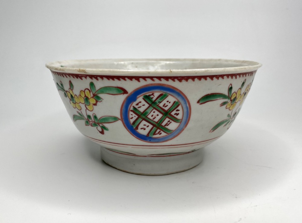 A Chinese Swatow porcelain bowl decorated with red, green, and yellow floral motifs and roundels on an off-white background. View V