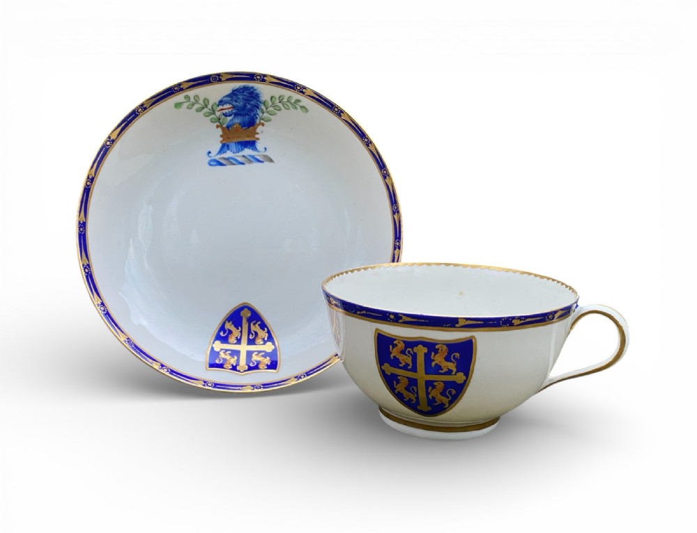 An 18th-century white porcelain teacup and saucer set decorated with cobalt blue rims, gilding, and heraldic coat of arms motifs.