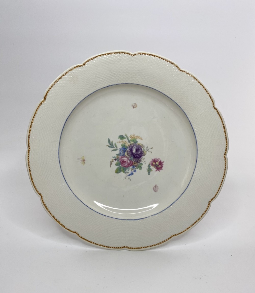 A circular off-white soft-paste porcelain plate with a scalloped edge, embossed basketweave rim, gilded border, and central hand-painted floral motif.