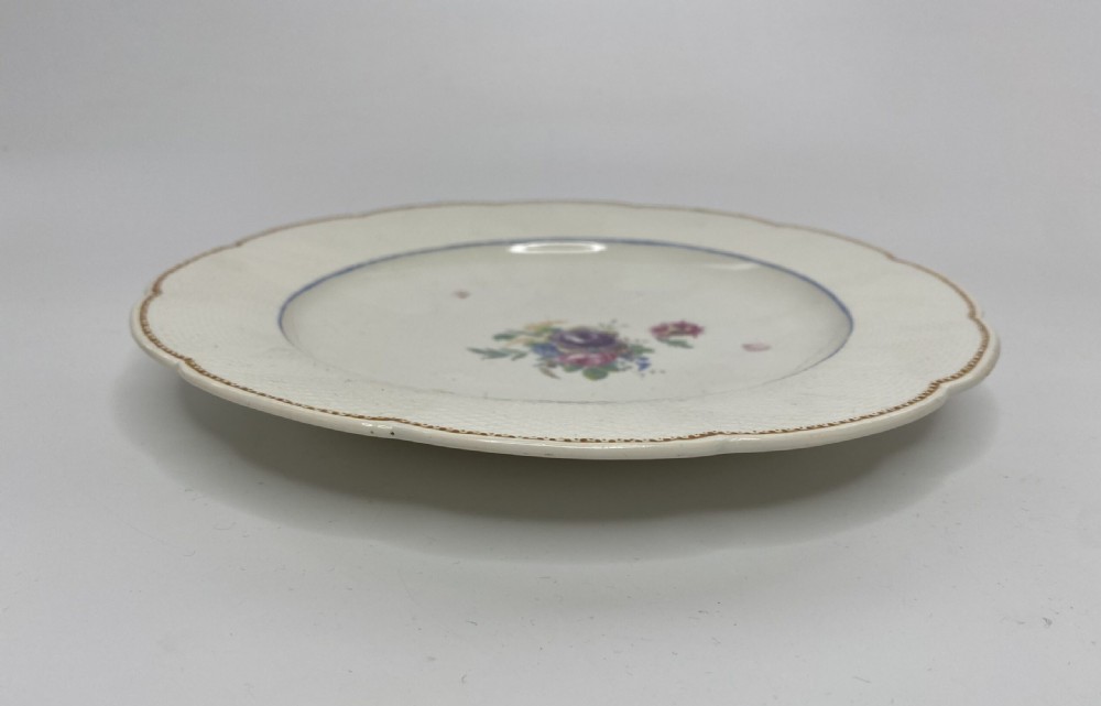 A circular off-white soft-paste porcelain plate with a scalloped edge, embossed basketweave rim, gilded border, and central hand-painted floral motif. View VI