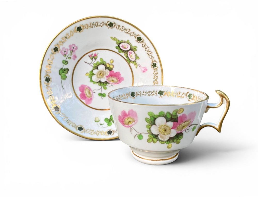 A Regency era white porcelain cup and saucer set decorated with hand-painted pink and white floral motifs and gilded rims.
