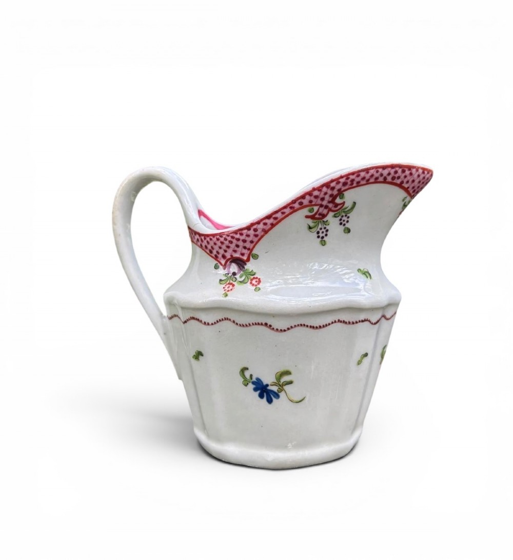 An 18th-century New Hall porcelain cream jug featuring hand-painted floral sprigs, a pink lattice border along the rim, and a faceted lower body with a high loop handle. View V