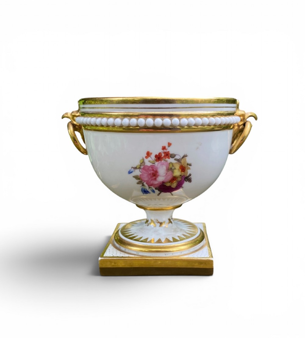 A Regency period white porcelain urn featuring hand-painted botanical blooms, twin gilt handles, a row of pearl-like beaded trimming around the rim, and a gilded square plinth base.