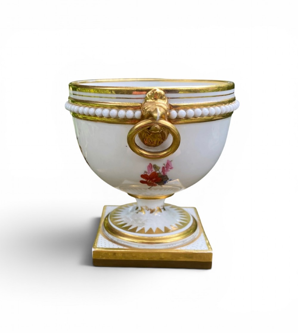 A Regency period white porcelain urn featuring hand-painted botanical blooms, twin gilt handles, a row of pearl-like beaded trimming around the rim, and a gilded square plinth base. View II
