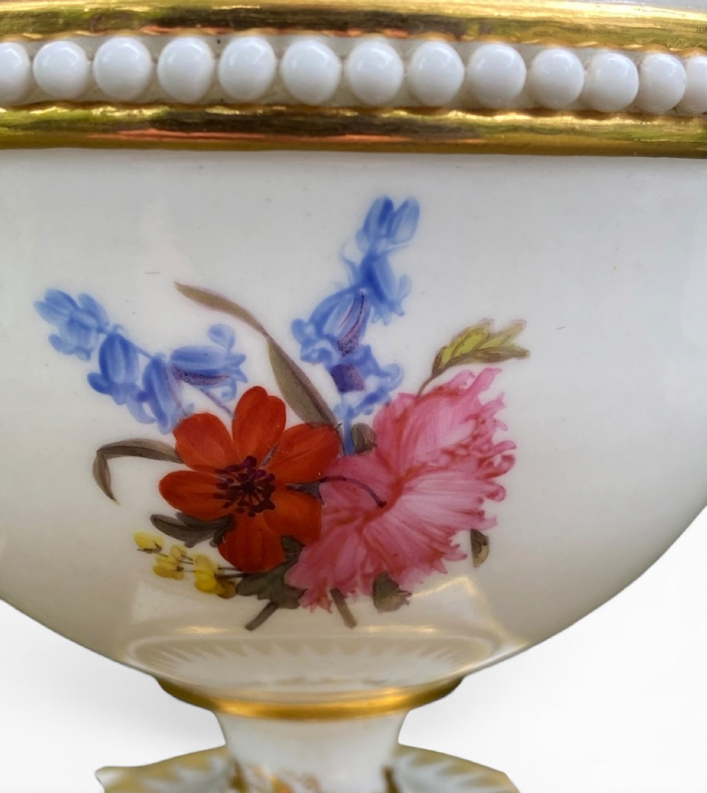 A Regency period white porcelain urn featuring hand-painted botanical blooms, twin gilt handles, a row of pearl-like beaded trimming around the rim, and a gilded square plinth base. View VII
