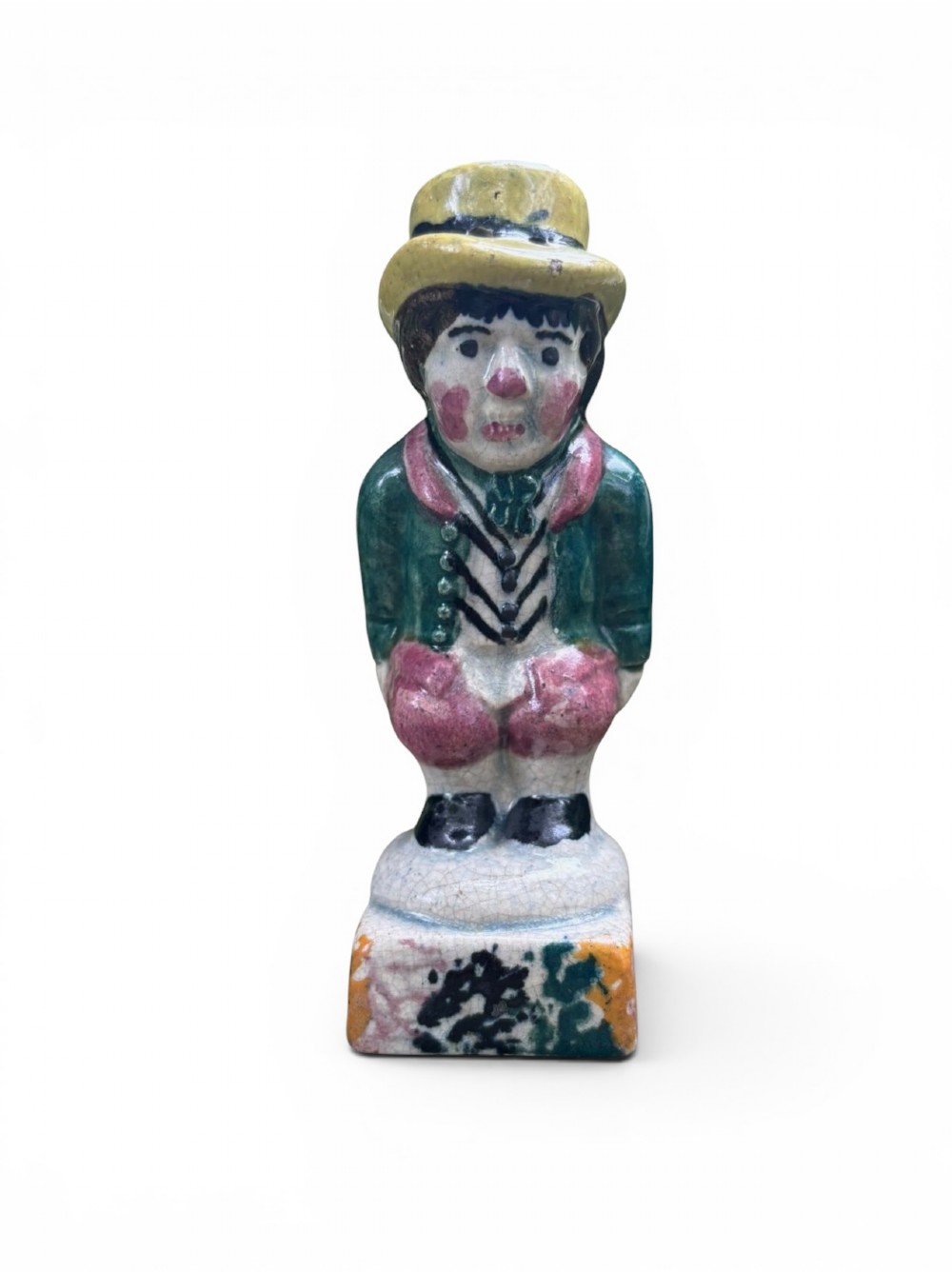 An antique ceramic Prattware figural pepper pot depicting a male character in a yellow top hat, green coat, and pink trousers on a square base.