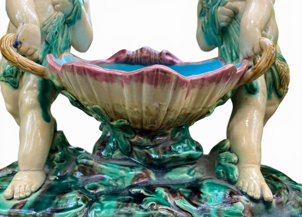 A Victorian Minton majolica centrepiece featuring two cherub figures supporting a shell-shaped bowl with a bright turquoise interior on a green foliate base. View VI