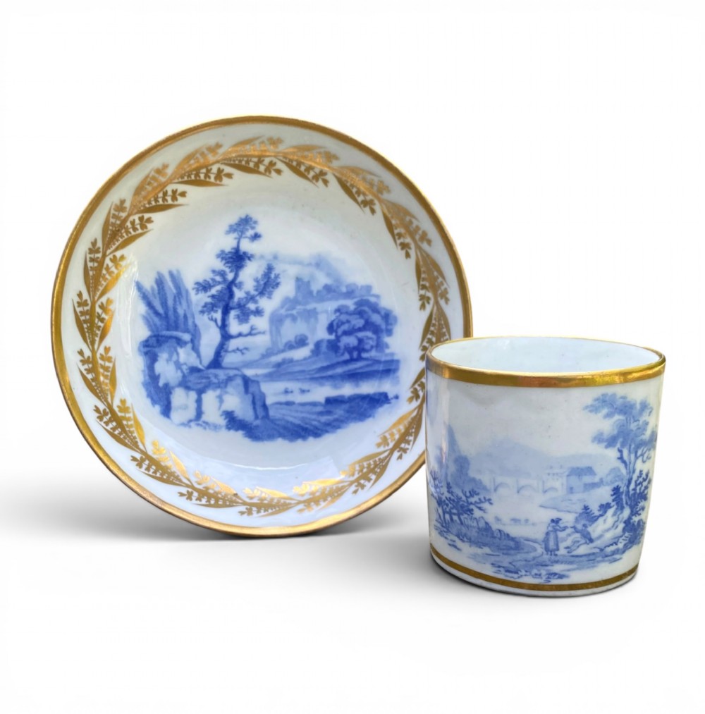 An antique blue and white porcelain coffee can and saucer featuring a painted landscape scene and gilded foliage borders.