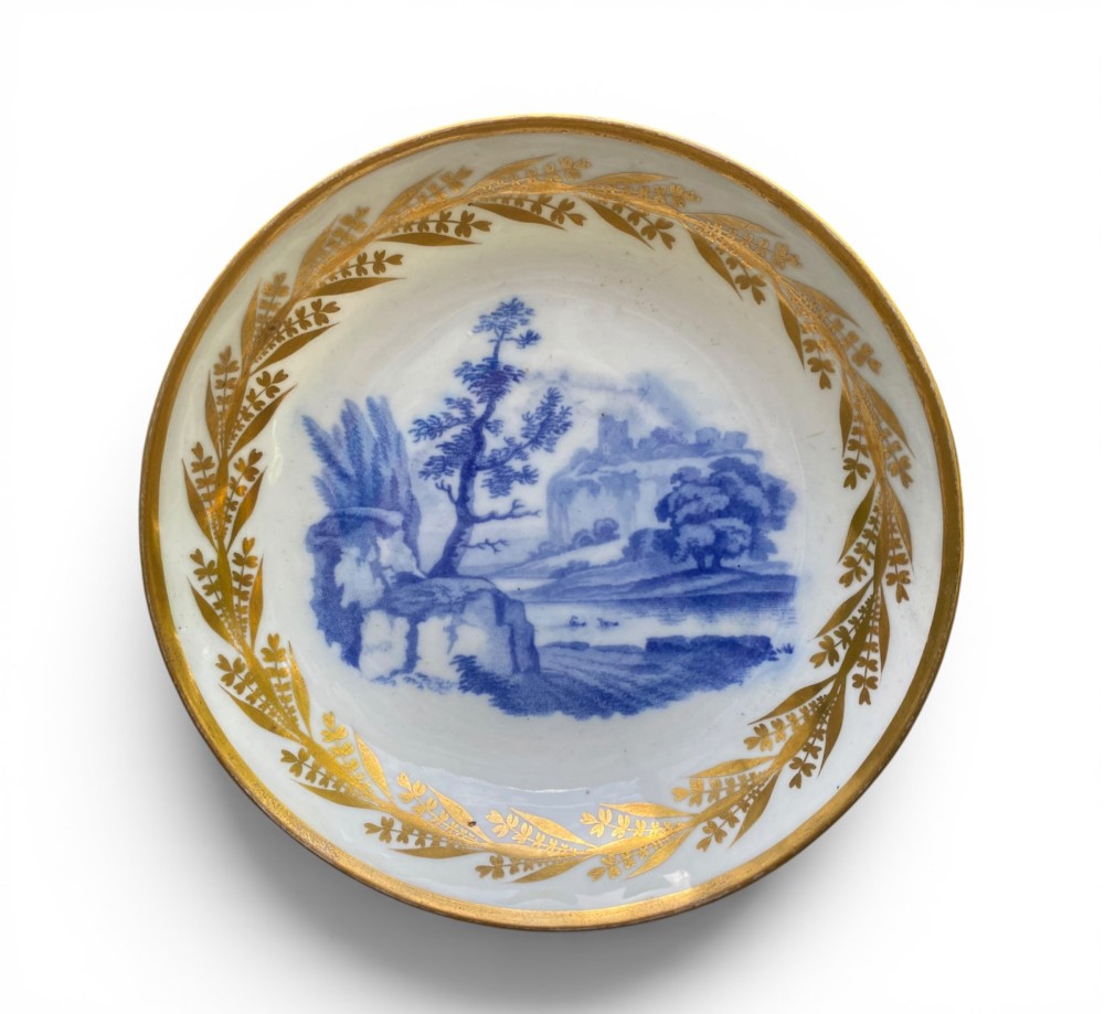 An antique blue and white porcelain coffee can and saucer featuring a painted landscape scene and gilded foliage borders. View II