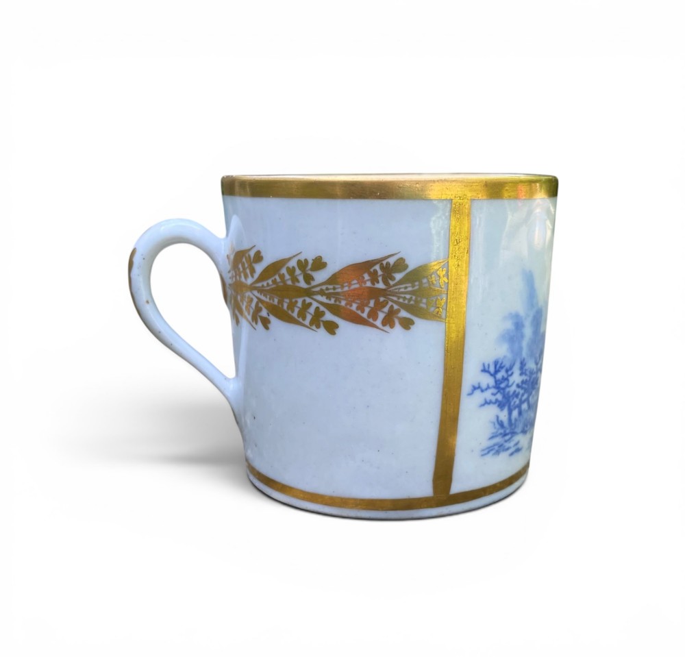 An antique blue and white porcelain coffee can and saucer featuring a painted landscape scene and gilded foliage borders. View VI