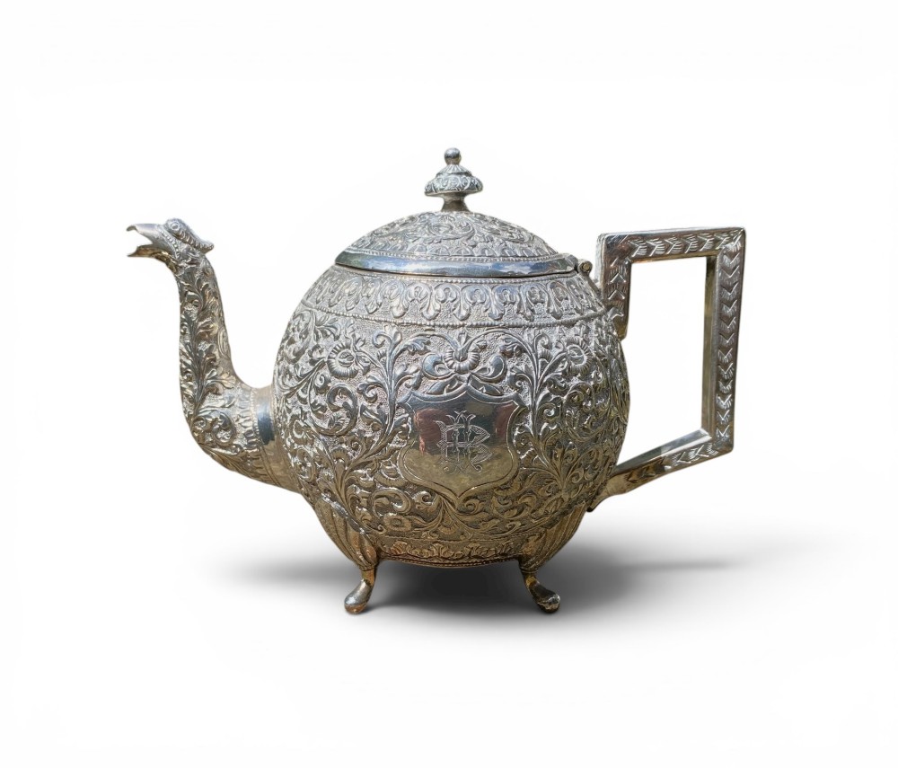 A three-piece antique Indian silver tea service featuring detailed chased floral scrollwork, elephant-shaped lid finials, and angular geometric handles. View IV