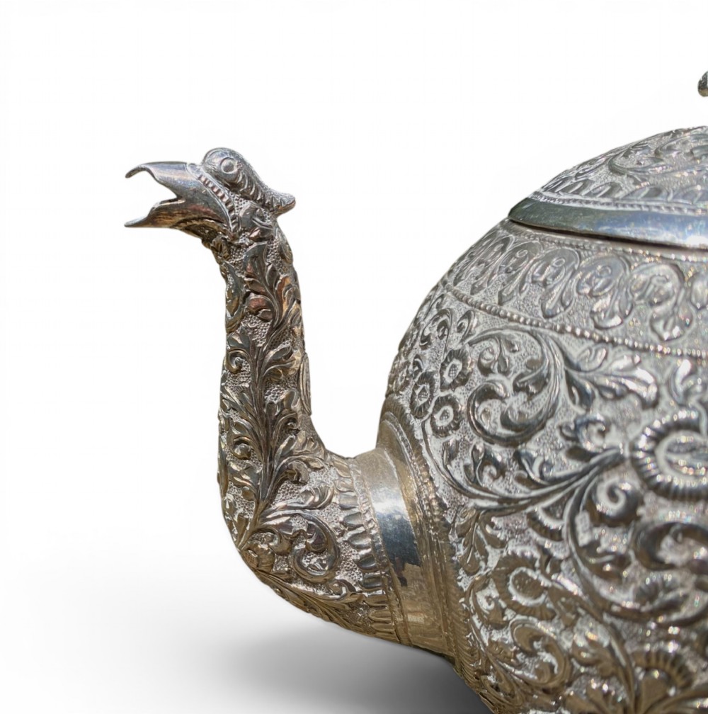 A three-piece antique Indian silver tea service featuring detailed chased floral scrollwork, elephant-shaped lid finials, and angular geometric handles. View V