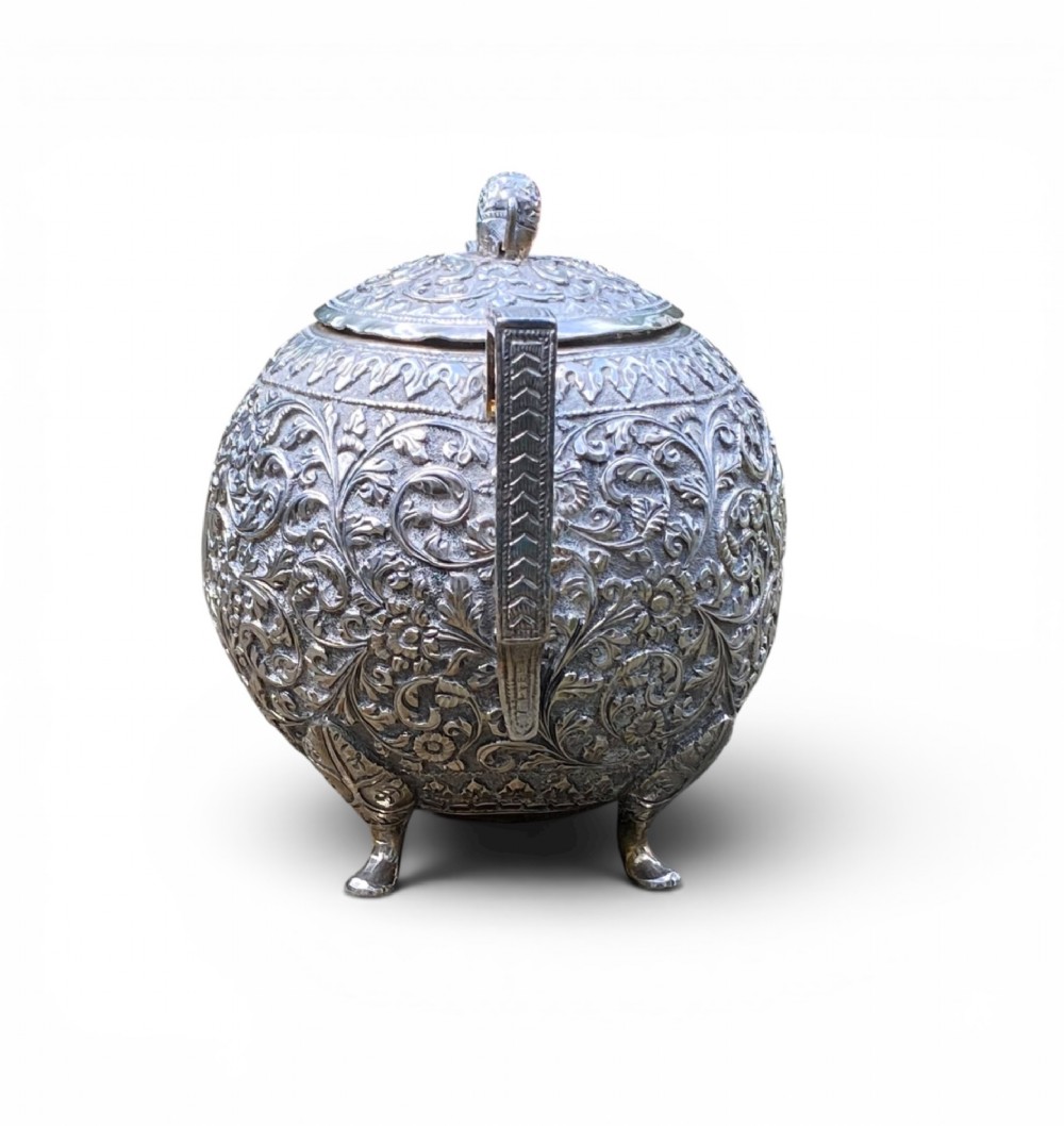 A three-piece antique Indian silver tea service featuring detailed chased floral scrollwork, elephant-shaped lid finials, and angular geometric handles. View IX