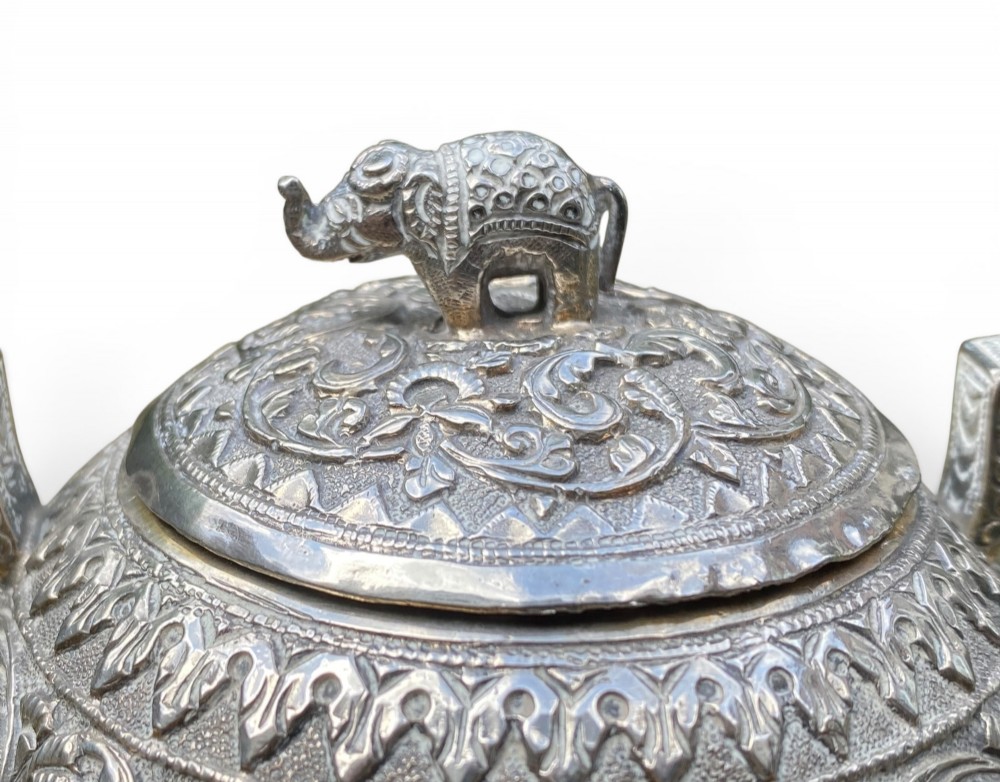 A three-piece antique Indian silver tea service featuring detailed chased floral scrollwork, elephant-shaped lid finials, and angular geometric handles. View X