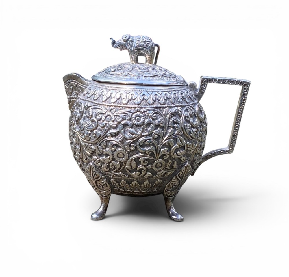 A three-piece antique Indian silver tea service featuring detailed chased floral scrollwork, elephant-shaped lid finials, and angular geometric handles. View XI
