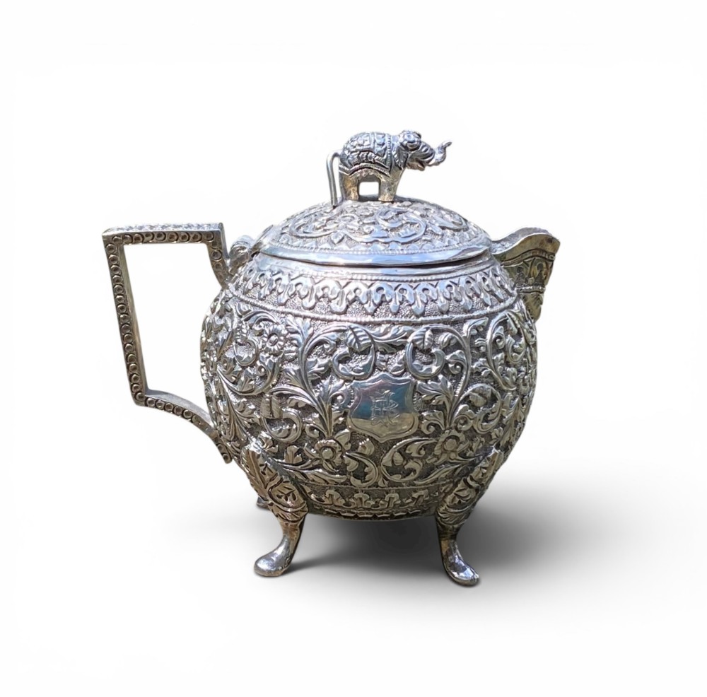 A three-piece antique Indian silver tea service featuring detailed chased floral scrollwork, elephant-shaped lid finials, and angular geometric handles. View XII