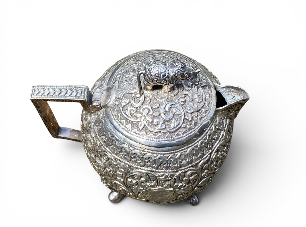A three-piece antique Indian silver tea service featuring detailed chased floral scrollwork, elephant-shaped lid finials, and angular geometric handles. View XIII