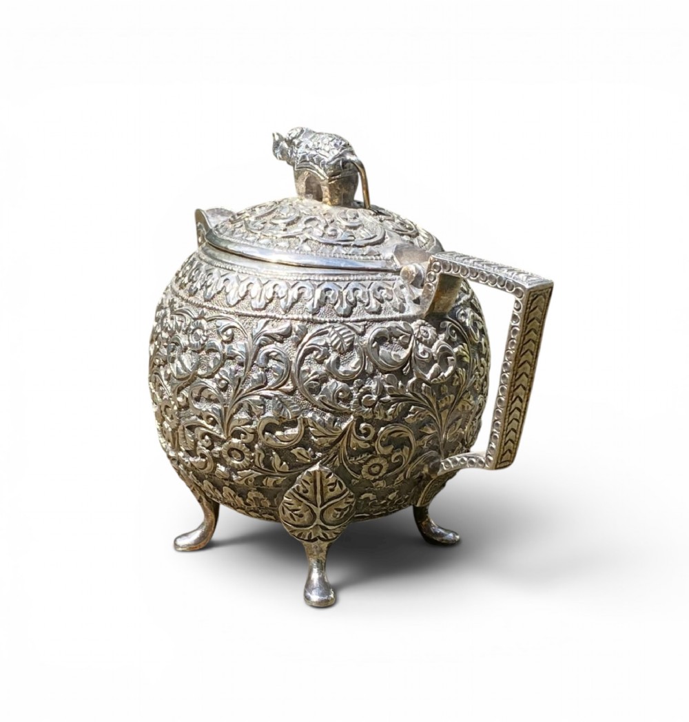 A three-piece antique Indian silver tea service featuring detailed chased floral scrollwork, elephant-shaped lid finials, and angular geometric handles. View XIV