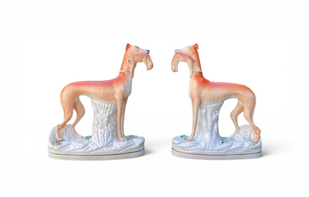A pair of Victorian Staffordshire earthenware greyhound figurines standing facing each other, each holding a hare in its mouth on a white ceramic base.