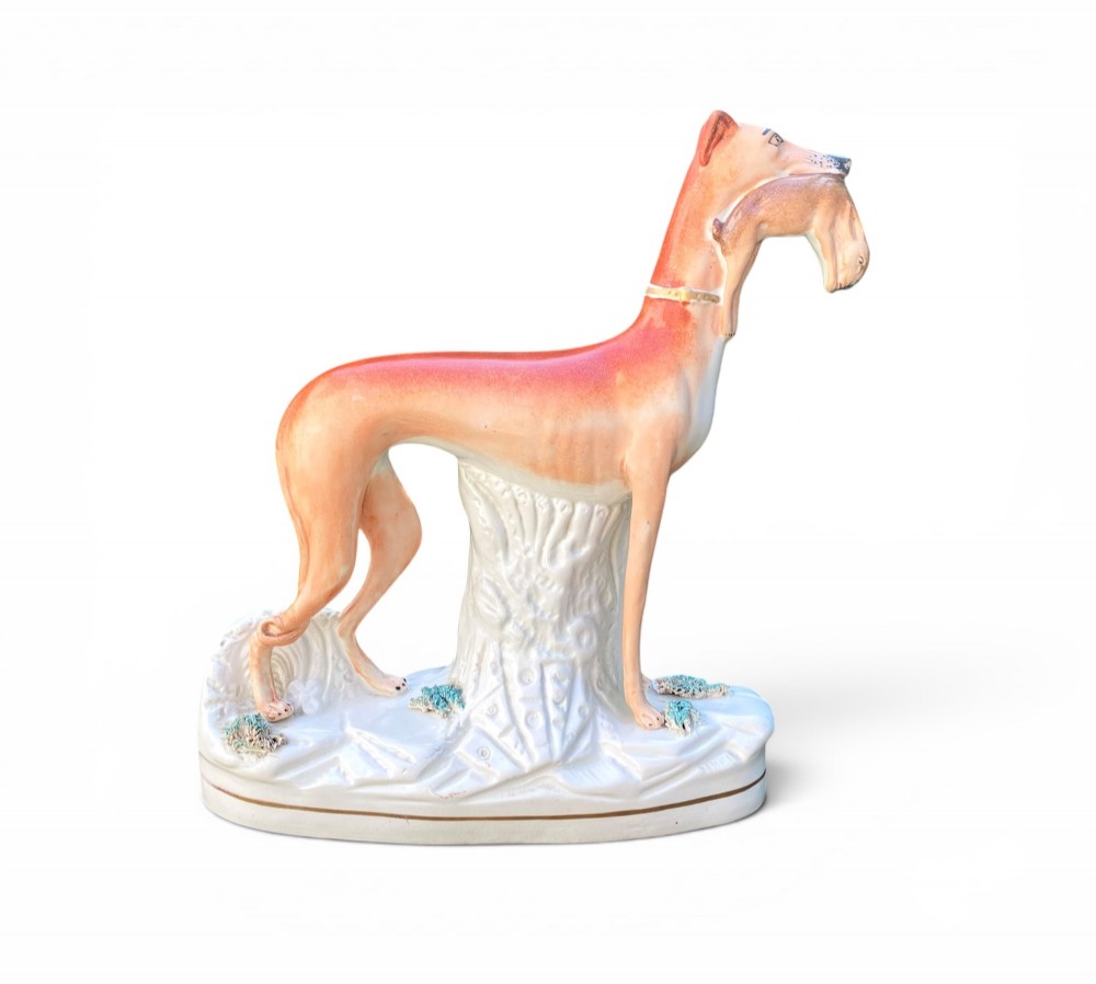 A pair of Victorian Staffordshire earthenware greyhound figurines standing facing each other, each holding a hare in its mouth on a white ceramic base. View II