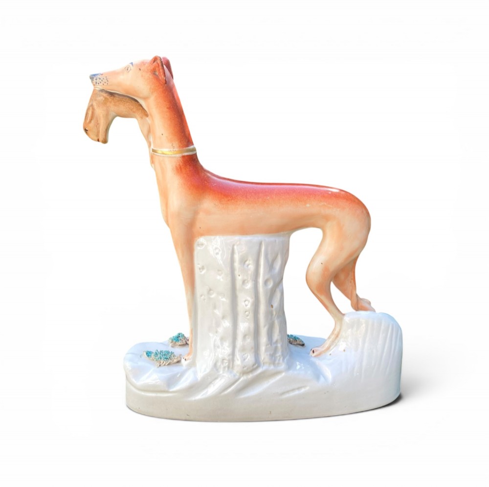 A pair of Victorian Staffordshire earthenware greyhound figurines standing facing each other, each holding a hare in its mouth on a white ceramic base. View IV