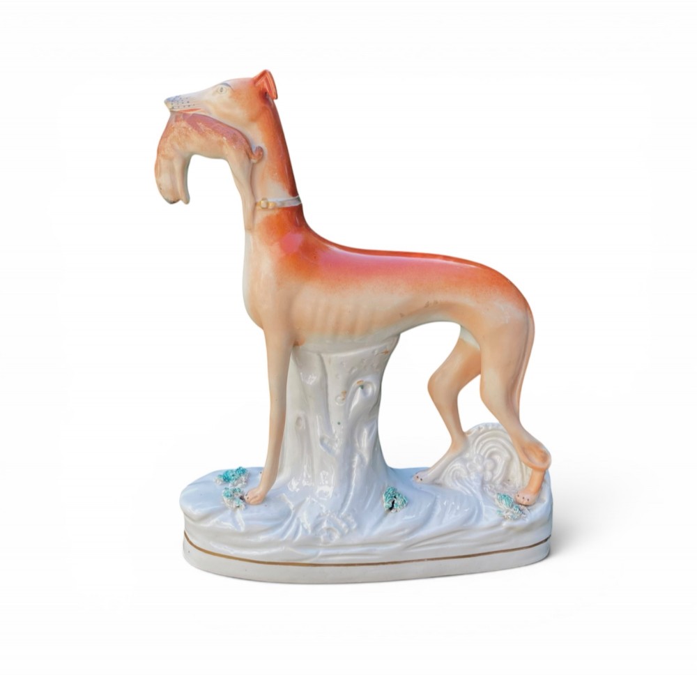 A pair of Victorian Staffordshire earthenware greyhound figurines standing facing each other, each holding a hare in its mouth on a white ceramic base. View VIII
