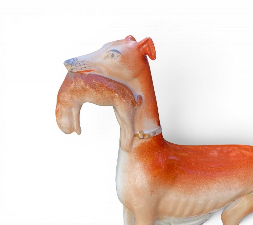 A pair of Victorian Staffordshire earthenware greyhound figurines standing facing each other, each holding a hare in its mouth on a white ceramic base. View IX