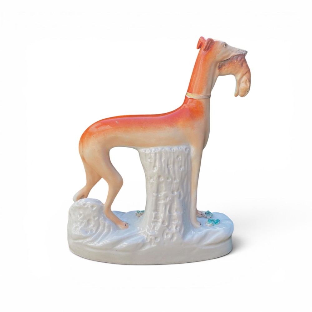 A pair of Victorian Staffordshire earthenware greyhound figurines standing facing each other, each holding a hare in its mouth on a white ceramic base. View X