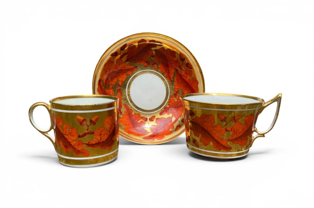 A Regency period Derby porcelain trio featuring two cups and a matching saucer decorated with red oak leaves and heavy gilding.