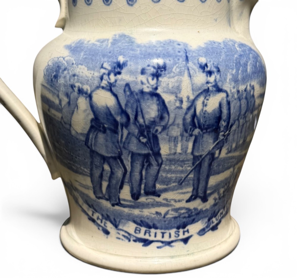 A cream earthenware jug featuring cobalt blue transferware depicting military figures labeled The British Volunteers. View III